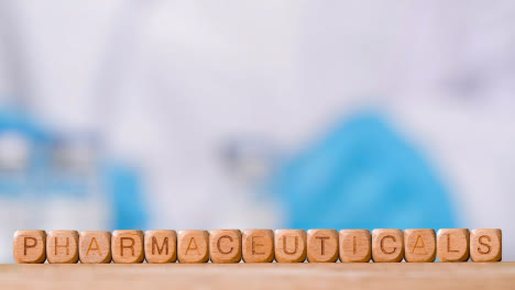 Medical-Concept-With-Wooden-Letter-Cubes-Or-Dice-Spelling-Pharmaceuticals-Against-Background-Of-Scientist-Working-In-Laboratory